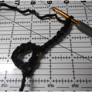 May include: Close-up of crochet work in progress. A black yarn is looped around a gold crochet hook with a gray handle. The yarn is on a white cutting mat with black grid lines.