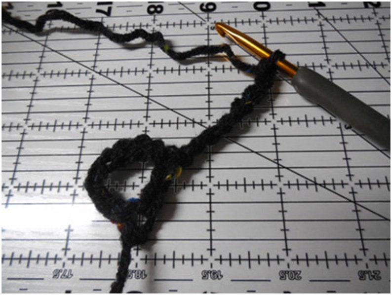 May include: Close-up of crochet work in progress. A black yarn is looped around a gold crochet hook with a gray handle. The yarn is on a white cutting mat with black grid lines.