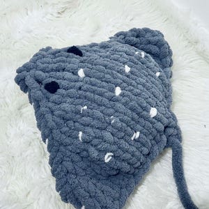 May include: A handmade, chunky knit hat in a soft, muted blue-grey colour. The hat features scattered white and black accents, adding a playful touch. The hat has a long, knitted tie. The hat is made of a thick, textured yarn, perfect for cold weather.