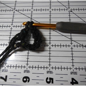 May include: Close-up of a crochet project in progress. A black yarn loop is held by a gold crochet hook with a gray handle. The project is on a white surface with black grid lines and measurements.