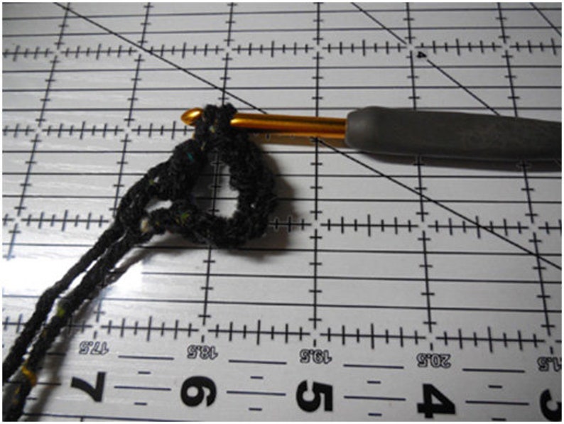 May include: Close-up of a crochet project in progress. A black yarn loop is held by a gold crochet hook with a gray handle. The project is on a white surface with black grid lines and measurements.