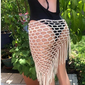 May include: A cream-colored, open-weave crochet skirt with a geometric pattern and fringe detail. The skirt is worn over a black bodysuit. The background features lush green foliage and potted plants, suggesting an outdoor setting.