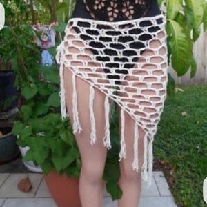 May include: A white, open-weave crochet skirt with a fringed hem. The skirt is asymmetrical, with a longer length on one side. The crochet pattern features large, open spaces, creating a see-through effect. The skirt is worn over a black garment.