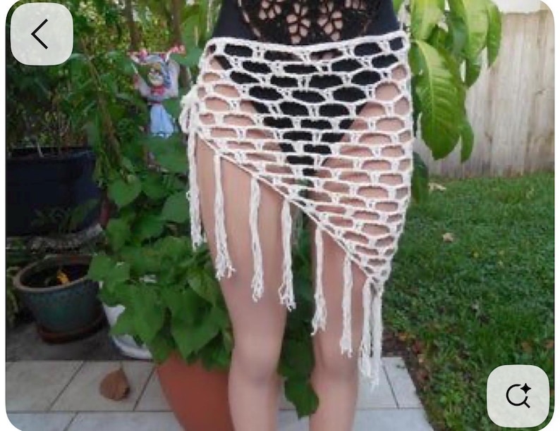 May include: A white, open-weave crochet skirt with a fringed hem. The skirt is asymmetrical, with a longer length on one side. The crochet pattern features large, open spaces, creating a see-through effect. The skirt is worn over a black garment.