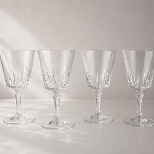 May include: A set of four clear glass wine glasses featuring a vertical cut pattern on the lower bowl and a faceted stem. Each glass has a wide rim, a tapered body, and a circular base, reflecting light through the transparent glass material.