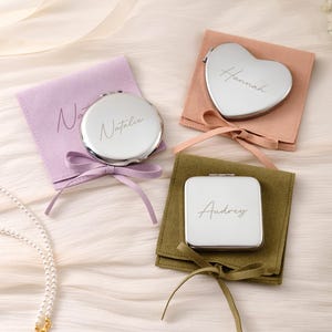 May include: Three silver-toned metal compact mirrors with personalized names engraved on the surface. One round mirror is on a lavender pouch, one heart-shaped mirror is on a peach pouch, and one square mirror is on an olive green pouch.