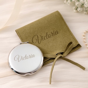 May include: A round silver-toned metal compact mirror featuring the name Victoria engraved on the front, accompanied by a matching olive green suede-like fabric pouch with a tie closure and the name Victoria embossed on the front surface.