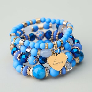 Peut inclure: Plusieurs bracelets de perles bleues superposés avec des détails dorés. Les bracelets présentent différentes nuances de bleu, dont le bleu clair, le bleu foncé et des perles transparentes. Un charm en forme de cœur doré avec le nom "Louisa" est suspendu.