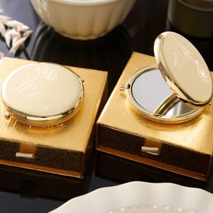 May include: Two gold compact mirrors, one open, rest on gold boxes. The compacts have floral designs and text. The boxes are square with a drawer. The background includes a white bowl and decorative elements.
