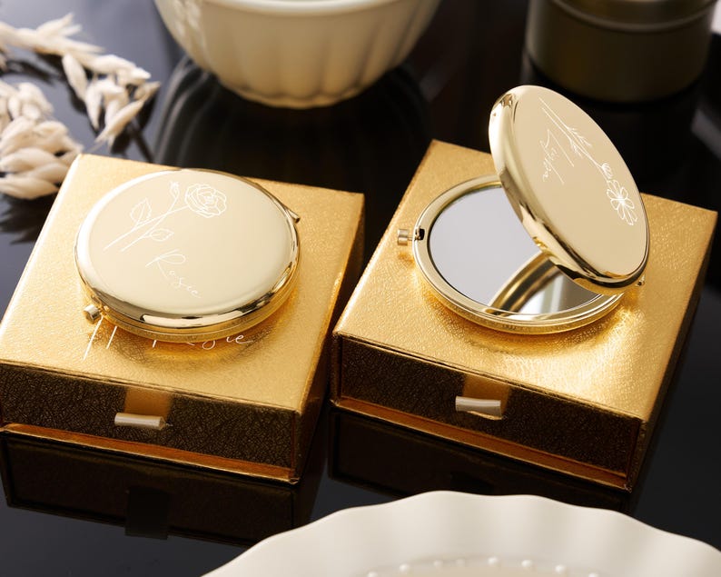 May include: Two gold compact mirrors, one open, rest on gold boxes. The compacts have floral designs and text. The boxes are square with a drawer. The background includes a white bowl and decorative elements.