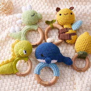 May include: A collection of handmade crochet baby rattles in various animal shapes, including a green dinosaur, yellow bee, blue whale, and yellow lemon. Each rattle features a wooden ring and is personalized with a name.