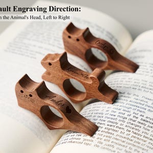 May include: Three wooden cat-shaped book holders with personalized names engraved on them, resting on an open book. The text "Default Engraving Direction: From the Animal's Head, Left to Right" is visible at the top. The book is open to page 174.
