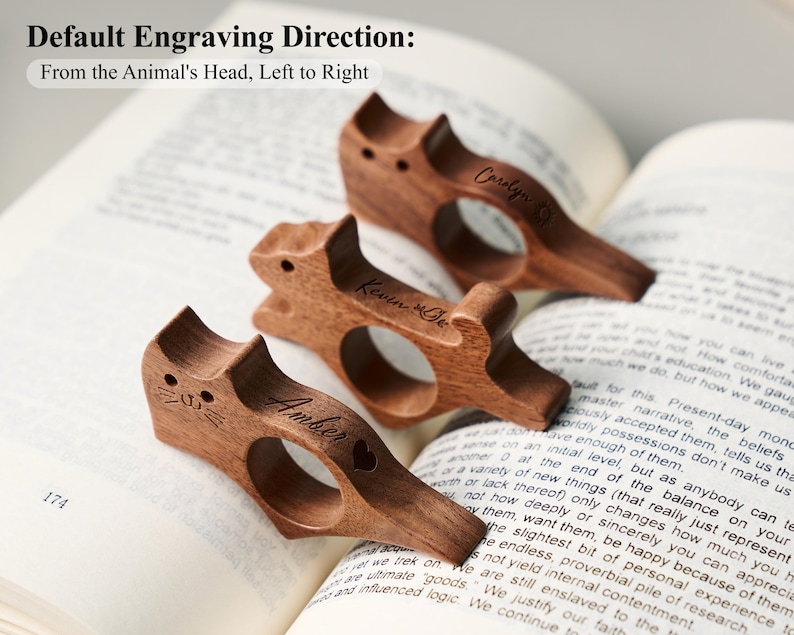 May include: Three wooden cat-shaped book holders with personalized names engraved on them, resting on an open book. The text "Default Engraving Direction: From the Animal's Head, Left to Right" is visible at the top. The book is open to page 174.