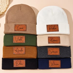 May include: Eight knitted beanies in a variety of colours, such as tan, white, green, and navy. Each beanie has a rectangular leather patch with a name written on it. The beanies are stacked on a light-coloured surface.