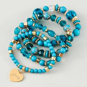 Peut inclure: Plusieurs bracelets de perles turquoise avec des détails dorés. L'un des bracelets comporte un charm en forme de cœur doré avec le nom "Jenny" inscrit. Les bracelets sont composés de perles de différentes formes et tailles, dont des perles rondes, ovales et facettées.