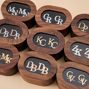 Op de afbeelding: Een verzameling houten sieradendoosjes met ovale vensters. Elke doos bevat een paar initialen in zilver of goud tegen een zwarte achtergrond. De dozen zijn in een groep gerangschikt en tonen verschillende initialencombinaties.