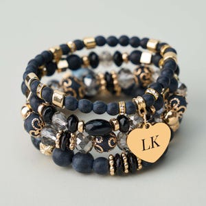 Puede incluir: Un conjunto de pulseras de cuentas en azul marino, negro y dorado. Las pulseras presentan diversas formas y tamaños de cuentas, incluyendo cristales facetados y un dije de corazón dorado con las iniciales "LK".