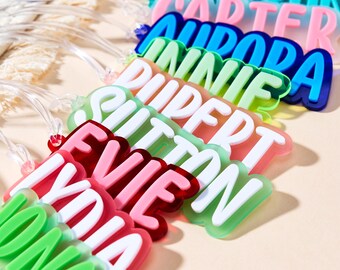 May include: A collection of colorful name tags in various shades, including pink, green, blue, and orange. Each tag features a different name in white, bold lettering. The tags are made of a glossy material and have clear plastic loops.
