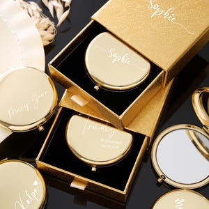 May include: Gold compact mirrors, some in open boxes, with personalized names like "Sophie", "Mary Jean", and "Trudy". The compacts are round or half-moon shaped, with a shiny finish. The boxes are gold with black interiors.