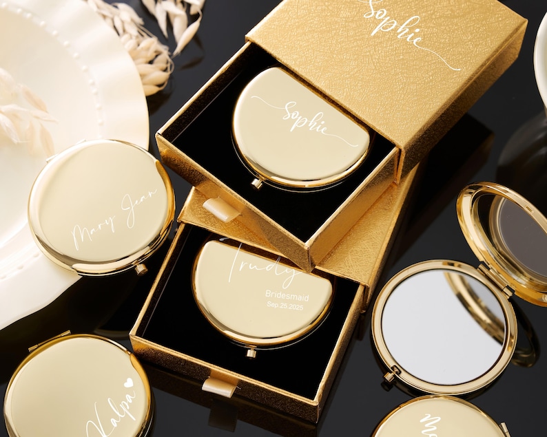 May include: Gold compact mirrors, some in open boxes, with personalized names like "Sophie", "Mary Jean", and "Trudy". The compacts are round or half-moon shaped, with a shiny finish. The boxes are gold with black interiors.