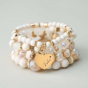 May include: A collection of ivory and gold beaded bracelets. The bracelets feature various beads, including round, faceted, and decorative beads. A gold heart charm with the name "Kadin" hangs from one bracelet. The bracelets are stacked together.