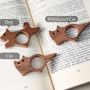 May include: Three wooden book page holders in the shape of animals: a dog, a cat, and a cat with whiskers. The holders are brown and sit on an open book. The text "Dog", "Cat", and "Whiskered Cat" are in speech bubbles.