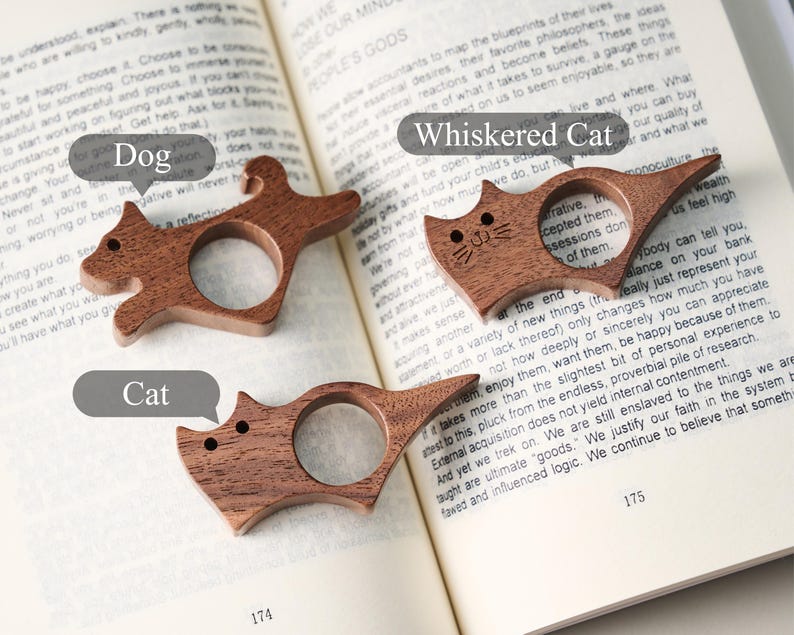 May include: Three wooden book page holders in the shape of animals: a dog, a cat, and a cat with whiskers. The holders are brown and sit on an open book. The text "Dog", "Cat", and "Whiskered Cat" are in speech bubbles.