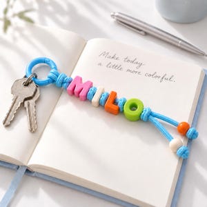 Könnte beinhalten: Ein Schlüsselanhänger mit einem blauen Kunststoffring, zwei silberfarbenen Metallschlüsseln und einer blauen Kordel, die mit bunten Buchstabenperlen den Namen MILO bildet und mit zwei kleinen runden Perlen am Ende versehen ist, auf einem Notizbuch.