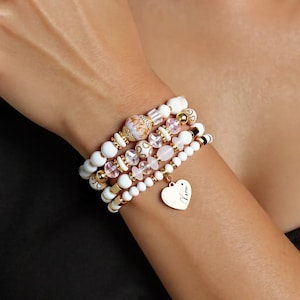 Op de afbeelding: Een stapel witte en gouden armbanden met een hartvormige bedel. De armbanden hebben verschillende kraalmaten, waaronder witte, gouden en roze kralen. De hartvormige bedel is goudkleurig en heeft de naam "Karen" gegraveerd.