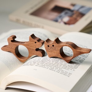 May include: Two wooden cat and dog-shaped book page holders. The dog-shaped holder has the name "Thomas" engraved on it, and the cat-shaped holder has the name "Ricarde" engraved on it. The holders are resting on the pages of an open book.