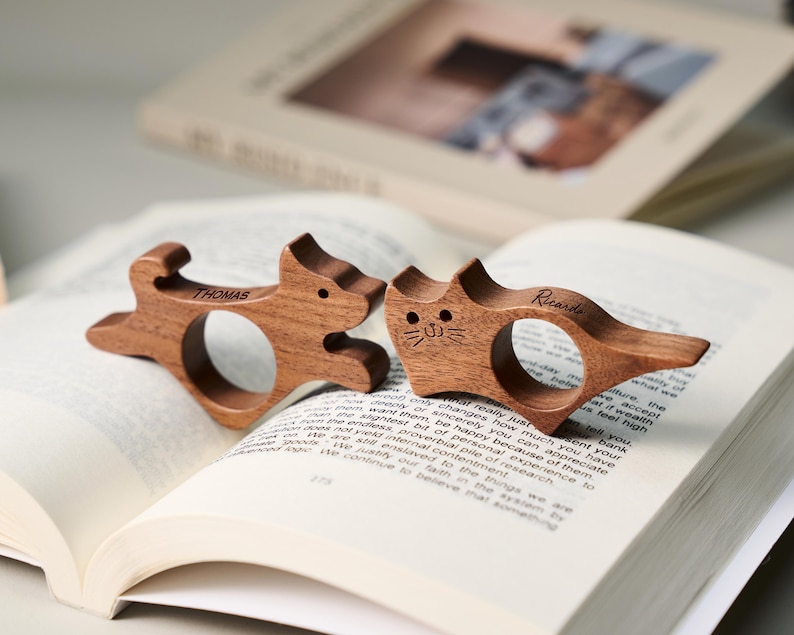 May include: Two wooden cat and dog-shaped book page holders. The dog-shaped holder has the name "Thomas" engraved on it, and the cat-shaped holder has the name "Ricarde" engraved on it. The holders are resting on the pages of an open book.