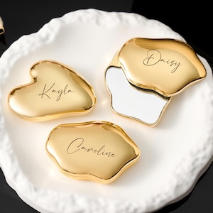May include: Three gold-colored trinket dishes with personalized names in cursive script. One is heart-shaped, another is an irregular shape, and the third is a petal shape. They sit on a white, organically shaped ceramic tray.