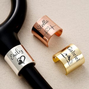 Pu&ograve; includere: Uno stetoscopio con tre fasce metalliche. Una fascia argentata con il nome "Emily" e un cuore. Una fascia color rame con il nome "Stella" e un grafico del battito cardiaco. Una fascia dorata con il nome "Gabriel" e una scena di montagna.