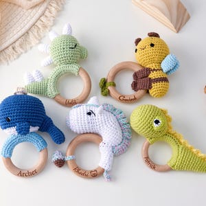 May include: Five crochet animal rattles attached to smooth wooden rings. The animals include a green dinosaur, a yellow bee, a blue whale, a white unicorn, and a light green dinosaur. Each wooden ring is engraved with a name: Violet, Govind, Archer, Rhodri, and Patrick.