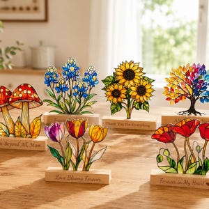 May include: Six decorative stained glass-style ornaments on wooden bases. Designs include red mushrooms, blue lupines, yellow sunflowers, a multicolored tree, tulips in purple, red, and yellow, and red poppies. Each base features engraved text.