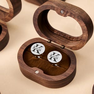 Può includere: Scatola ovale in legno con coperchio incernierato, aperta per rivelare un paio di gemelli in argento. Ogni gemello ha una faccia bianca con la lettera "K" e il nome "Kevin" stampato in nero. La scatola è foderata in velluto marrone.