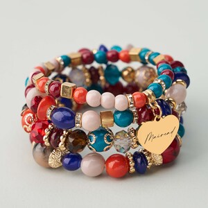 Può includere: Un gruppo di braccialetti di perline colorate, tra cui rosso, blu, arancione e rosa. I braccialetti presentano un mix di perline rotonde e quadrate, con un ciondolo a forma di cuore dorato con la scritta "Mairead".