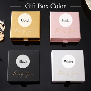 May include: Four square gift boxes in gold, pink, black, and white. Each box has a circular label indicating its color and the name "Mary Jean" in cursive. The boxes are arranged on a dark surface.