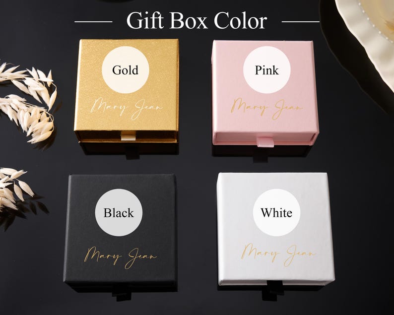 May include: Four square gift boxes in gold, pink, black, and white. Each box has a circular label indicating its color and the name "Mary Jean" in cursive. The boxes are arranged on a dark surface.