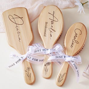 May include: Three wooden hairbrushes with personalized names and floral designs. Each brush is tied with a white ribbon that reads "Just For You." One brush is rectangular, while the other two are oval-shaped. A perfume bottle is visible in the background.