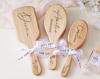 May include: Three wooden hairbrushes with personalized names and floral designs. Each brush is tied with a white ribbon that reads "Just For You." One brush is rectangular, while the other two are oval-shaped. A perfume bottle is visible in the background.