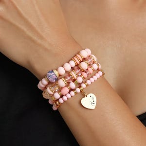 Peut inclure: Plusieurs bracelets de perles roses avec des détails dorés et un charm en forme de cœur. Les bracelets présentent différentes tailles et textures de perles, y compris des perles transparentes et à motifs. Le charm en forme de cœur est gravé d'un nom.
