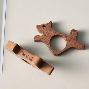 May include: Two wooden dog-shaped napkin rings. One is in the shape of a running dog, and the other is a rectangular shape with the name "David" engraved on it. Both are made of a light brown wood.