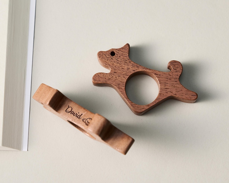 May include: Two wooden dog-shaped napkin rings. One is in the shape of a running dog, and the other is a rectangular shape with the name "David" engraved on it. Both are made of a light brown wood.