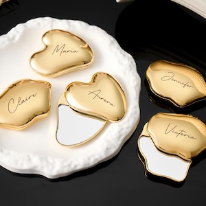 May include: Gold-colored Gua Sha tools with personalized names, including "Maria", "Claire", "Aurora", "Jennifer", and "Victoria". The tools are displayed on a white, organically shaped plate against a black background.