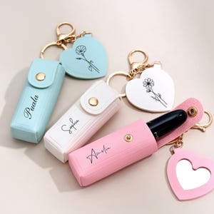 May include: Three rectangular lipstick cases in light blue, white, and pink, each featuring a gold snap closure and a personalized name. Attached to each case is a gold keyring and a heart-shaped charm, with one pink heart featuring a mirror.