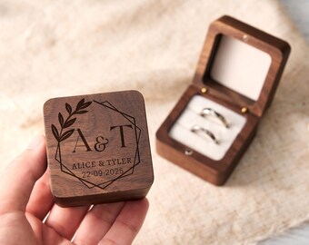 May include: A wooden ring box with a geometric design and the initials "A & T" engraved on the lid. The box is open, revealing two rings inside. The date "22-09-2025" is also engraved on the box.