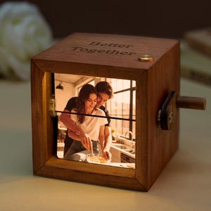 Op de afbeelding: Een houten muziekdoos met een foto van een stel erin. De doos is kubusvormig met de woorden "Better Together" gegraveerd op de bovenkant. Een kleine slinger en een messingkleurige knop zijn zichtbaar. De foto toont een stel in een keuken.