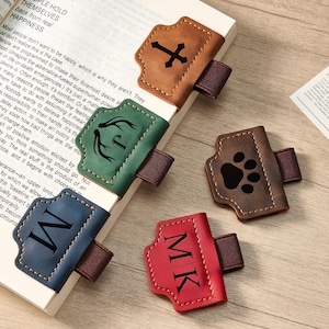 May include: Leather book page holders in various colors: brown with a cross, green with antlers, blue with a letter "W", brown with a paw print, and red with the initials "MK". Each has an elastic band.