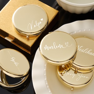 May include: Gold-colored compact mirrors with personalized names in white script. One mirror is open, revealing a reflective surface. The mirrors are on a white plate and a gold gift box. The names include "Kalpa", "Sophie", "Marlene", "Veslihan", and "Mary Jean".